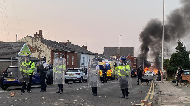 Smoke billows from a fire started by protesters as riot police stand guard after disturbances near the Southport Islamic Society Mosque in Southport, northwest England, on July 30, 2024.