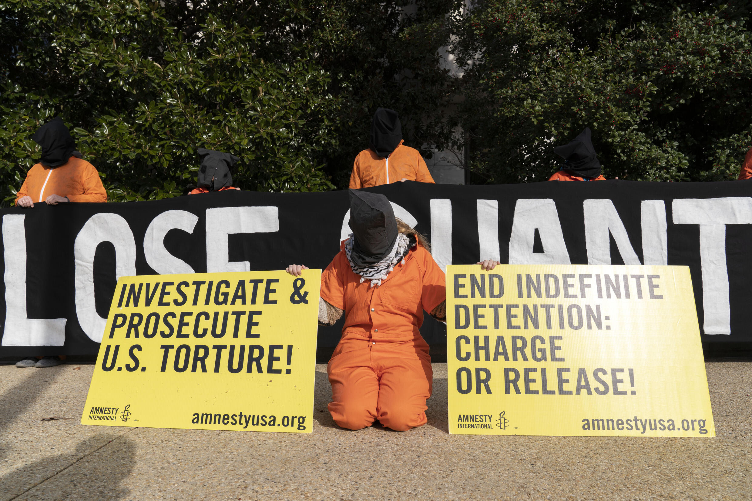 Des manifestants portant des combinaisons de prisonniers orange et une cagoule noire, symbolisant les détenus de Guantanamo, tiennent une banderole demandant la fermeture de la prison sur la colline du Capitole à Washington, le mardi 7 décembre 2021.