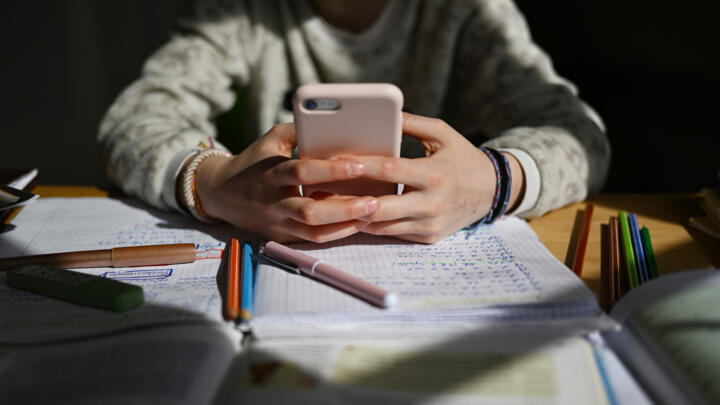 This illustration photo shows a schoolchild sitting at a desk with school materials and holding a smartphone in their hands.