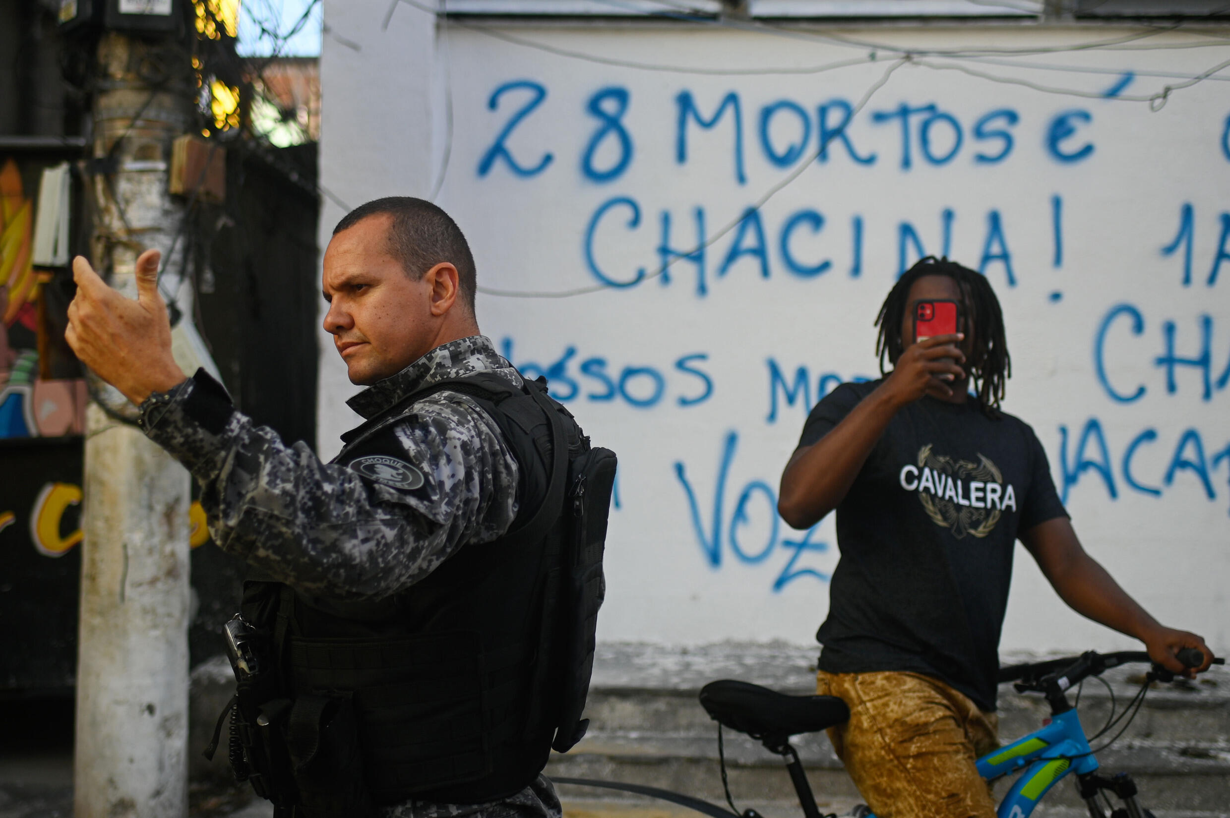 A resident records a video with his cell phone as an officer monitors a protest against police violence in the favela of Jacarezinho on May 6, 2022, the first anniversary of the deadliest police raid in Rio de Janeiro history
