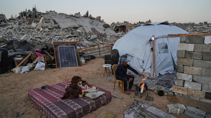 A displaced Palestinian sits by a fire outside his tent amid the destruction left by an Israeli offensive in Gaza City, on October 22, 2025.