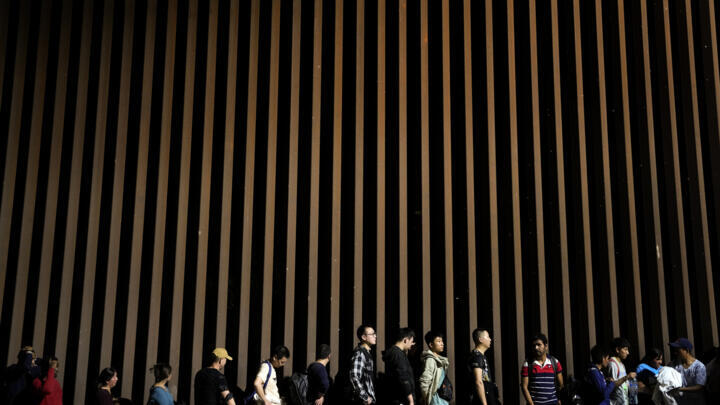 People line up against a border wall as they wait to apply for asylum after crossing the border from Mexico, Tuesday, July 11, 2023, near Yuma, Ariz.